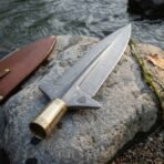 Damascus spearhead with winged lugs and runed brass socket lying on a river stone beside a brown leather sheath