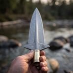 Damascus spearhead with winged lugs, engraved valknut and runed brass socket held up in a hand by a river