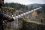 Damascus steel arming sword with a brown leather grip and brazil nut pommel held out by a man on a ruined stone wall, wooded hills behind