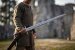Damascus steel arming sword with knotwork panel above the guard held at the side by a man near castle ruins