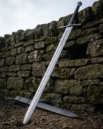 Damascus steel bastard sword with long black grip and straight cross leaning on a dry stone wall above its black scabbard