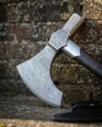 Damascus steel bearded axe head with engraved eye leaning against an old brick wall with its sheath at the base