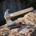 Damascus steel bearded axe standing on a rock ledge beside its brown leather blade sheath, helm of awe etched cheek, brass collar and ash haft