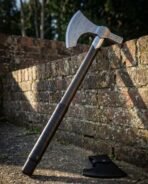 Damascus steel bearded axe with engraved hammer poll and dark haft leaning on a brick wall above a black sheath