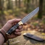 Damascus steel broken back seax with a horn handle, brass bolster and rune etched blade held in hand, leather sheath on a log behind