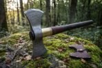 Damascus steel double bit axe with a dark hardwood haft, brass collar and leather grip wrap, resting on a mossy rock beside its leather sheath