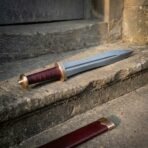 Damascus steel gladius with a leaf shaped blade, bronze meander guard and oxblood leather grip on stone steps beside its scabbard