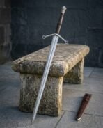 Damascus steel hand and a half sword propped against a weathered stone bench with its brown leather scabbard nearby