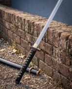 Damascus steel katana with black ito wrapped tsuka and pale rayskin under the wrap leaning on an old brick wall, saya on the gravel