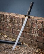 Damascus steel katana blade leaning at an angle against a mossy brick wall with the black lacquered saya laid at its foot