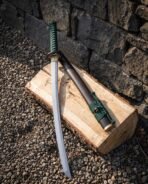 Damascus steel katana with green cord wrapped handle leaning on a log beside its dark wooden saya against a stone wall