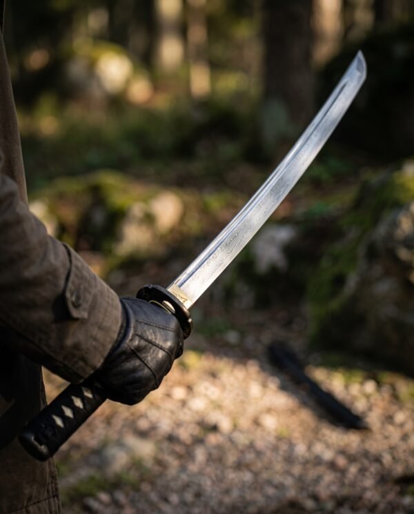 Damascus steel katana with visible pattern and temper line held in a gloved hand, black wrapped handle in a forest