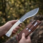 Damascus steel kukri knife with bone handle scales and wolf etched steel pommel held in hand, brown leather sheath holding two small companion knives