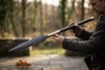 Damascus steel leaf blade spear with a rune cut brass socket and dark wooden shaft held across the body by a man outdoors