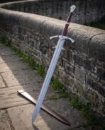 Damascus steel longsword with a barbed guard, crown pommel and oxblood grip leaning on a stone bridge wall by its scabbard