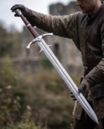 Damascus steel longsword with barbed guard held point down in gloved hands by a man before castle ruins