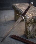 Damascus steel longsword with faceted pommel and quatrefoil guard leaning on a stone bench above its leather scabbard