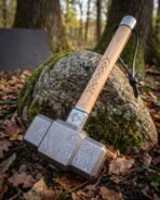 Damascus steel Mjolnir style hammer with a carved knotwork ash handle and steel end cap, propped against a mossy boulder among fallen leaves