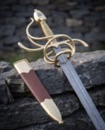 Damascus steel rapier with a brass swept hilt and wire wrapped grip resting on a dry stone wall beside its brass mounted scabbard