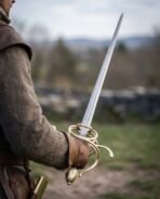 Damascus steel side sword with a brass toned swept hilt and knuckle bow held out over a green valley by a man in wool
