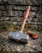 Damascus steel sledge hammer resting head down on a stone ledge against a dry stone wall, leather cover beside it