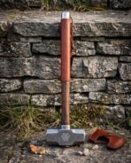 Damascus steel sledge hammer stood upright on its head against a stone wall with the brown leather cover at its side