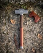 Damascus steel sledge hammer with valknut head and leather wrapped wood handle laid on stone beside a leather head cover
