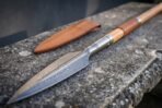 Damascus steel spear with leaf blade, steel socket and leather wrapped ash haft lying on stone beside a tan leather head sheath
