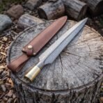Damascus steel spearhead with brass socket collar laid diagonally beside its brown leather sheath on a stump