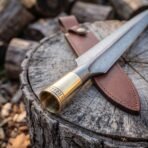 Damascus steel spearhead with rune engraved brass socket collar lying on a tree stump beside a brown leather sheath
