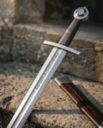Close up of a Damascus steel sword blade with an engraved rune row, steel cross guard and wheel pommel on a stone step beside the scabbard