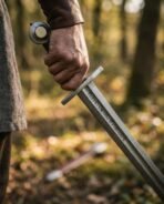 Damascus steel sword with a wheel pommel and rune row on the blade gripped in a bare hand, timber scabbard on the forest floor behind