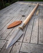 Damascus steel Viking spear with leaf blade, brass collar and ash shaft lying on wooden decking beside a leather cover