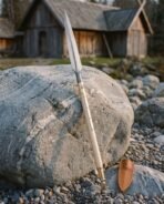 Damascus steel Viking spear with leaf head and pale ash haft leaning on a boulder in front of timber longhouses
