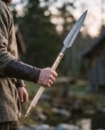 Damascus steel Viking spear with leaf shaped head and brass fittings held in a hand beside a wooden building