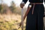 Damascus steel Viking sword held point down beside a man in black with its brown leather scabbard hanging from his belt