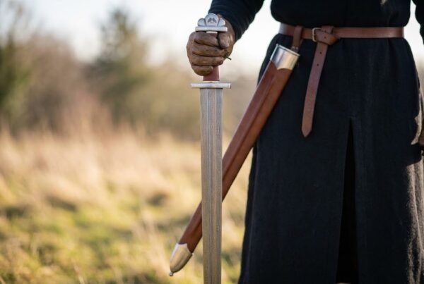Damascus steel Viking sword held point down beside a man in black with its brown leather scabbard hanging from his belt
