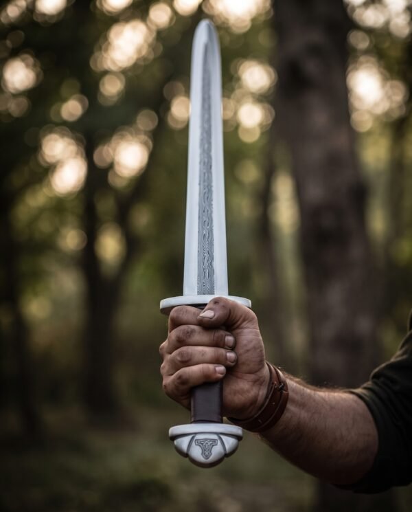 Damascus steel Viking sword held upright in a fist against blurred forest light, pommel and brown grip in view