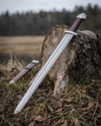 Damascus steel Viking sword leaning on a tree stump in a field with its brown leather scabbard in the grass