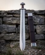 Damascus steel Viking sword stood beside its brown leather scabbard with steel chape against a stone wall
