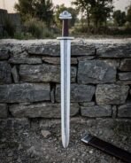 Damascus steel Viking sword stood point down in front of a dry stone wall, scabbard on the gravel below