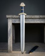 Damascus steel Viking sword with brass guard and engraved disc pommel standing against a grey wooden bench