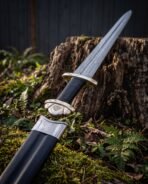 Damascus steel Viking sword with brass three lobed pommel and black grip resting on its black scabbard by a tree stump