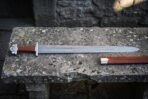 Damascus steel Viking sword with five lobed steel pommel and brown leather grip laid on old stone beside its leather scabbard