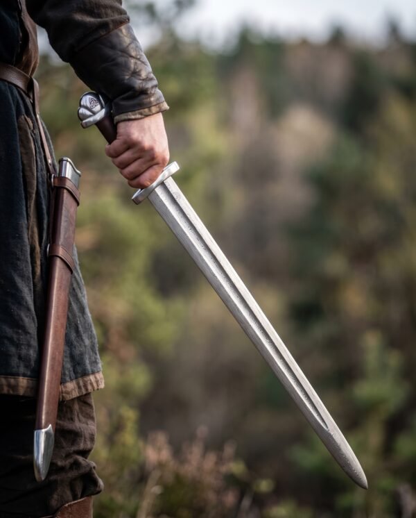 Damascus steel Viking sword with a fullered pattern blade and lobed pommel held at the side beside a brown leather scabbard