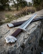 Damascus steel Viking sword with valknut on the lobed pommel lying across a stone wall with its brown leather scabbard