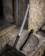Damascus steel wakizashi with black cord wrapped tsuka leaning on a stone doorway beside its black lacquer saya