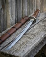 Damascus steel wakizashi with brown leather wrapped handle lying on weathered wood beside its brown leather saya