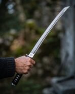 Damascus steel wakizashi with Celtic knot collar and black cord wrapped grip held up in one hand against a dark forest
