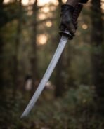 Damascus steel wakizashi with curved blade held in a gloved hand in a dark forest, knotwork tsuba in view